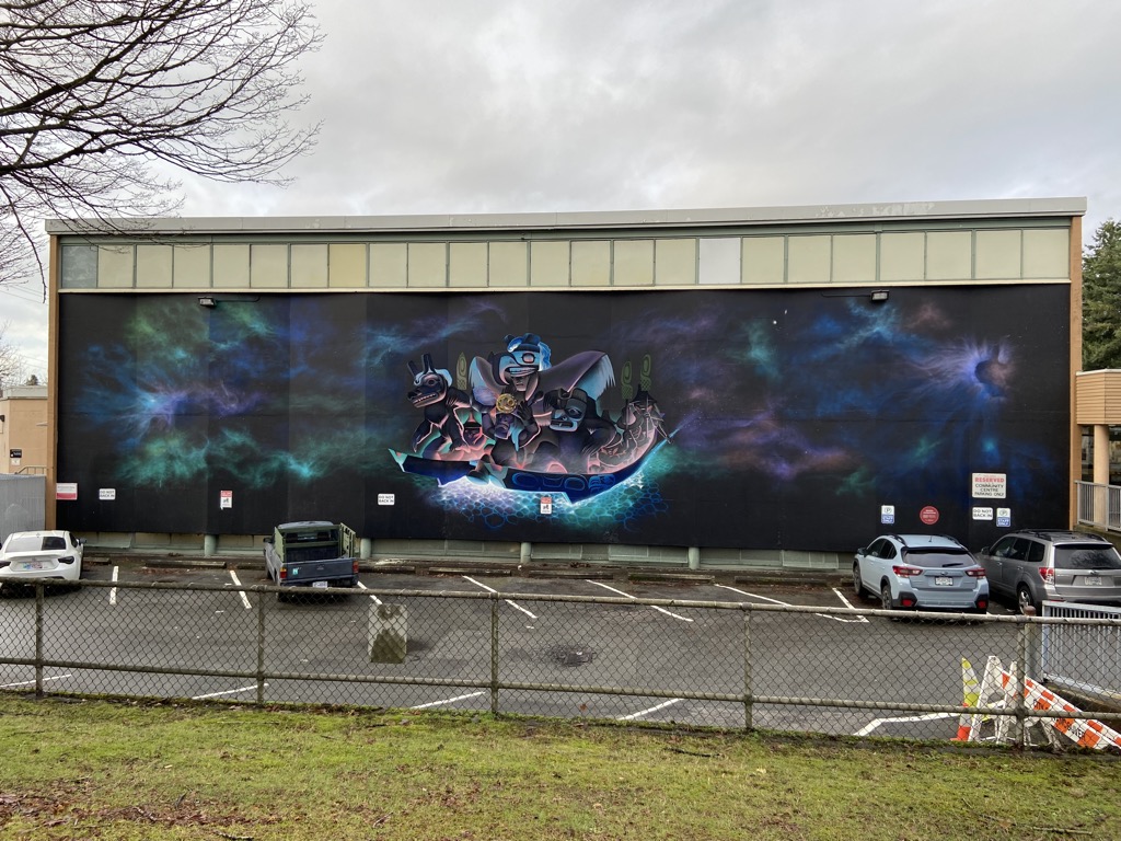 view of "The Future has Arrived / Aatl'aan K'atgwaa Tluu. K'aatlaagang," a large-scale mural painted on the dark exterior wall of the Hastings Community Centre in Vancouver, BC. The mural depicts Northwest Coast Indigenous figures in a glowing canoe set against a cosmic, aurora-like background. The mural spans the full width of the building above the Centre's parking lot.