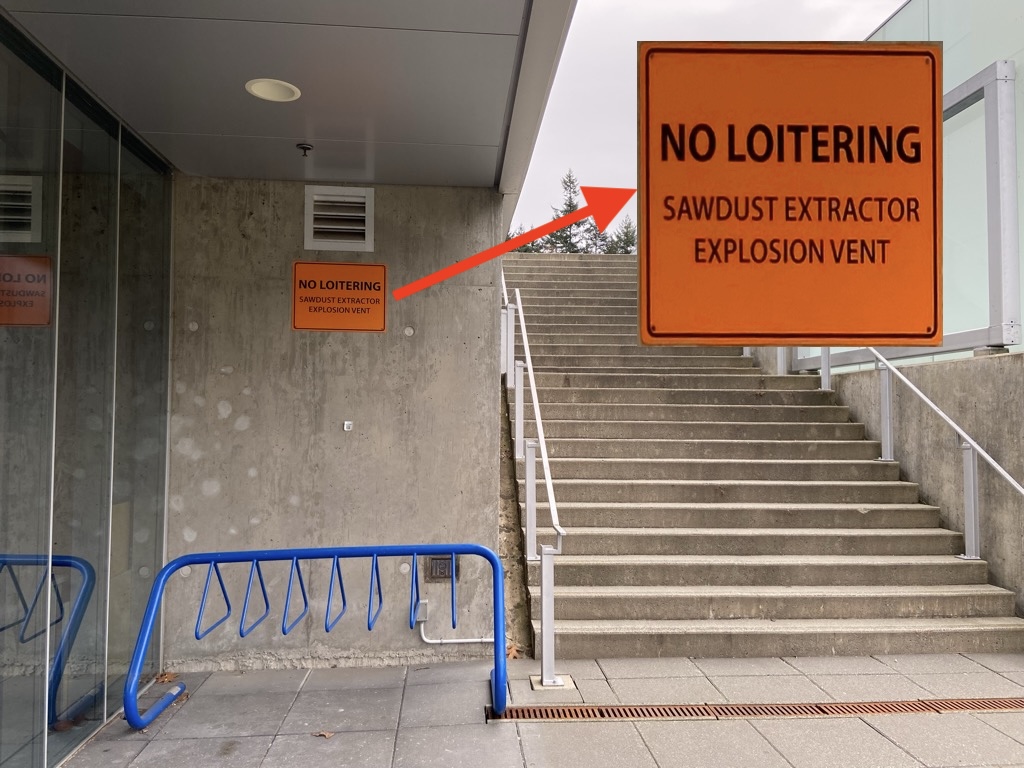 Concrete stairwell entrance with a blue bike rack on the left and an orange safety sign on the wall reading 'No Loitering - Sawdust Extractor Explosion Vent.'