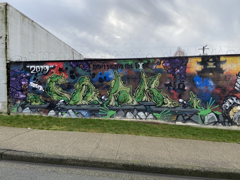 Left portion of a large graffiti mural on a concrete block wall in Vancouver. Features large green blob-like characters with toothy grins sitting on a ledge, surrounded by colorful splashes of red, blue, and purple. Text reads '2019' and 'Don't Believe the Hype.' Purple cartoon villain characters float in the upper corners.