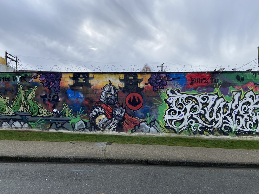 Center portion of a large graffiti mural. A heavily armored knight or samurai figure in grey armor with a red cape and horned helmet stands prominently, bearing a circular red emblem. Purple flying characters with weapons appear above. To the right, large ornate white wildstyle graffiti lettering begins. Text reads 'Bring the...' Japanese-style castle architecture is visible in the background.