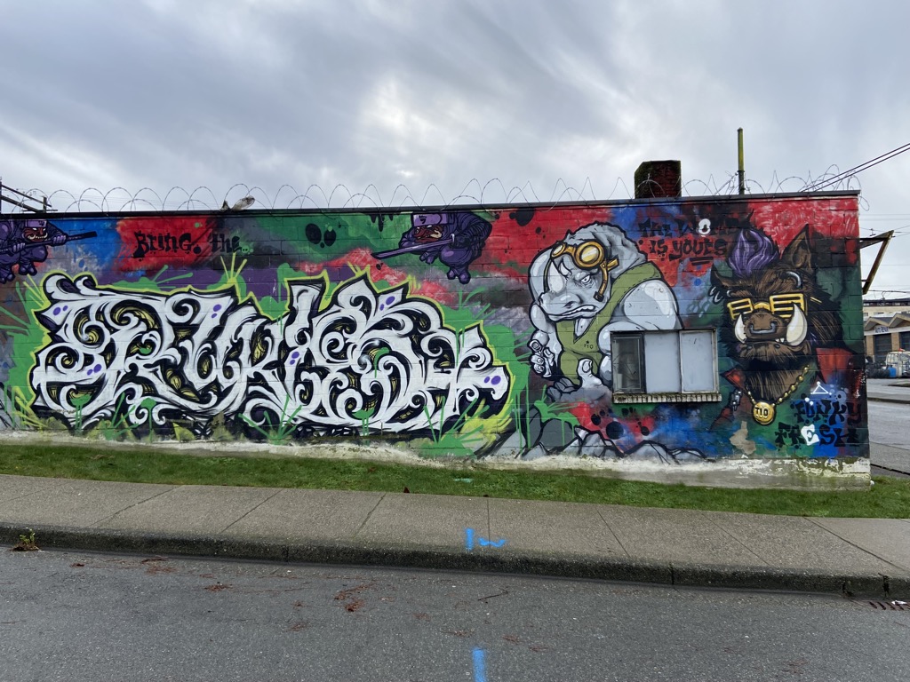 Right portion of a large graffiti mural wrapping around a building corner. Dominant white and gold wildstyle graffiti lettering fills the left half. To the right, two cartoon animal characters: a grey rhino wearing goggles and a brown wolf/bear wearing gold sunglasses and a chain medallion. Text reads 'Bring the...' and 'The World Is Yours.'