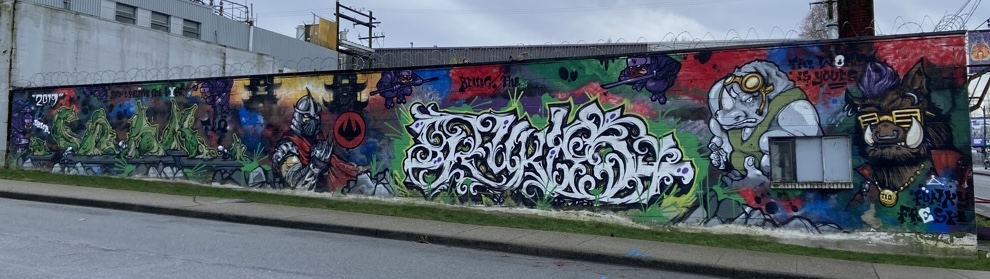 Wide-angle view of the complete graffiti mural spanning the exterior wall of Storm Brewing in Vancouver's East Side. The mural depicts green blob creatures, a medieval knight, large wildstyle lettering, and cartoon animal characters, set against vibrant red, blue, green, and yellow backgrounds. Text across the mural reads '2019,' 'Don't Believe the Hype,' and 'Bring the...'